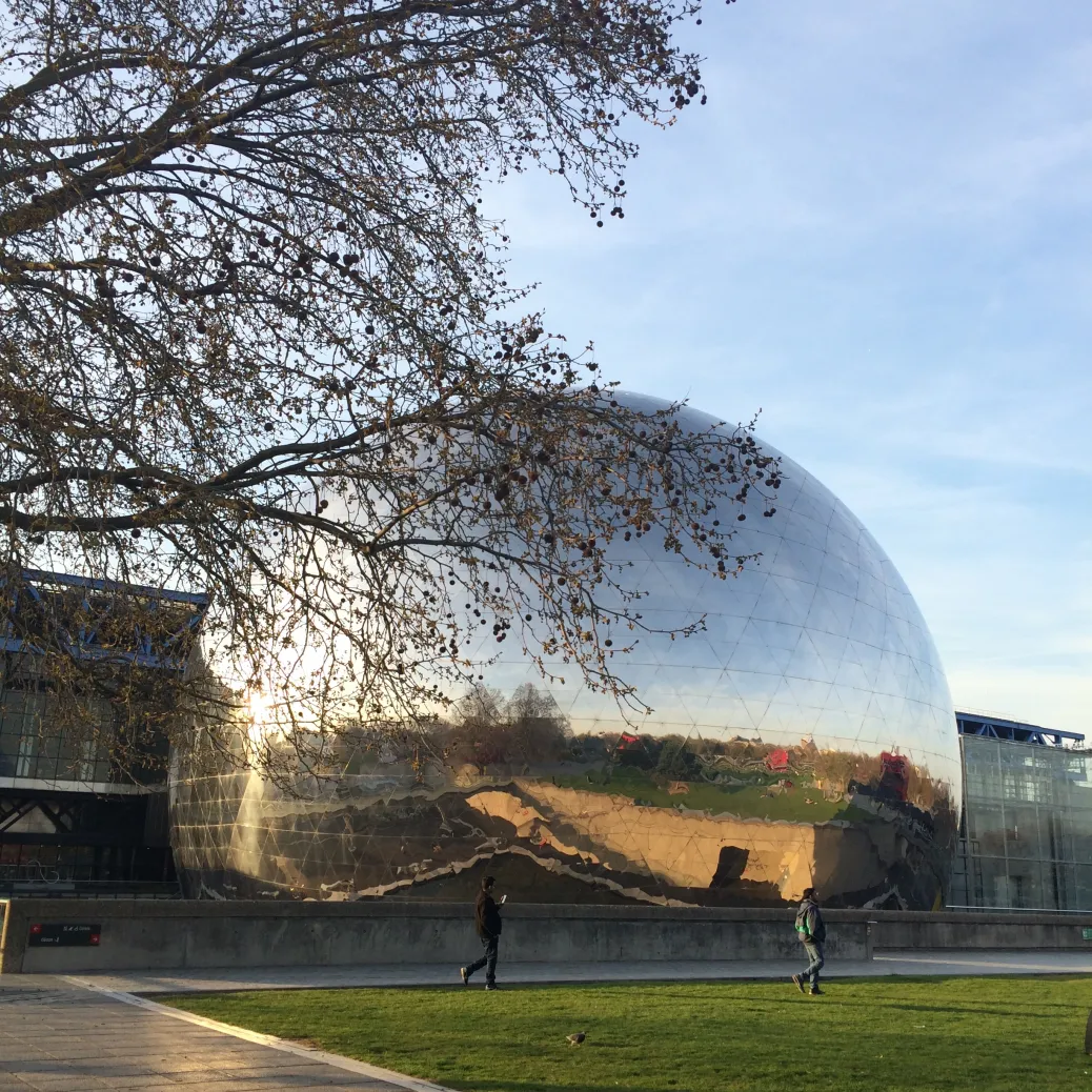 Photographie argentique au grain visible d'une grande sphère miroir reflétant la verdure et le ciel, avec des branches d'arbres nues au premier plan