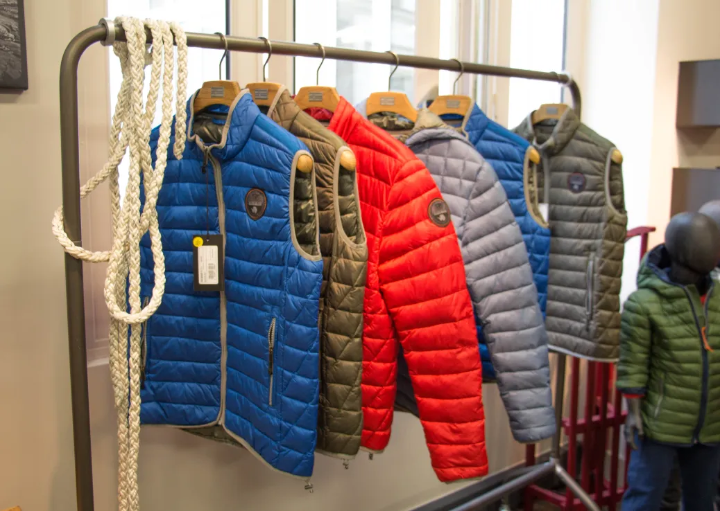 Étagère de vestes matelassées colorées en bleu, vert, rouge et gris près d'une fenêtre, lumière douce, ambiance chaleureuse.